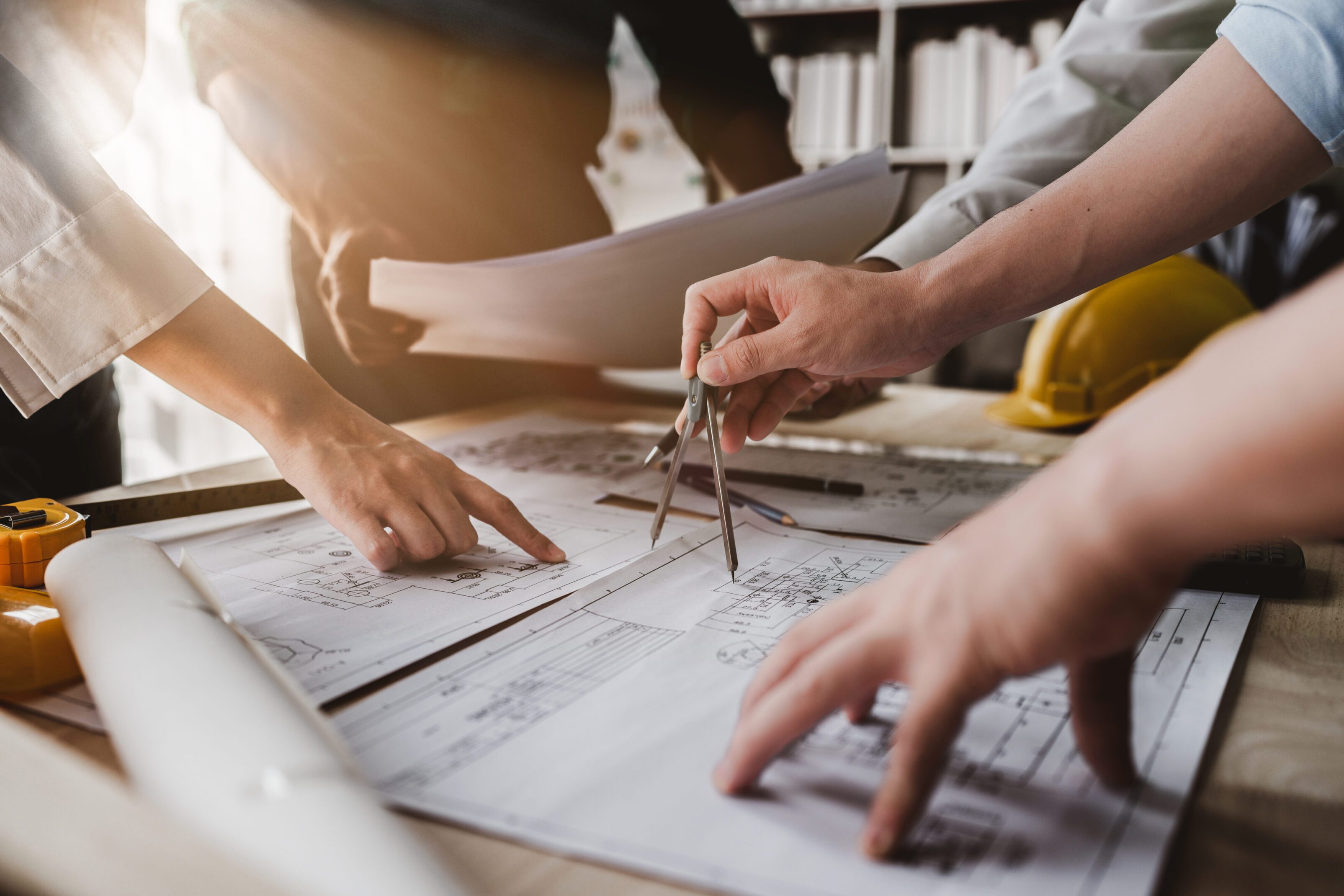 Three people review and discuss architectural blueprints on a table, one holding a compass and others pointing at the plans; a yellow hard hat is visible in the background.