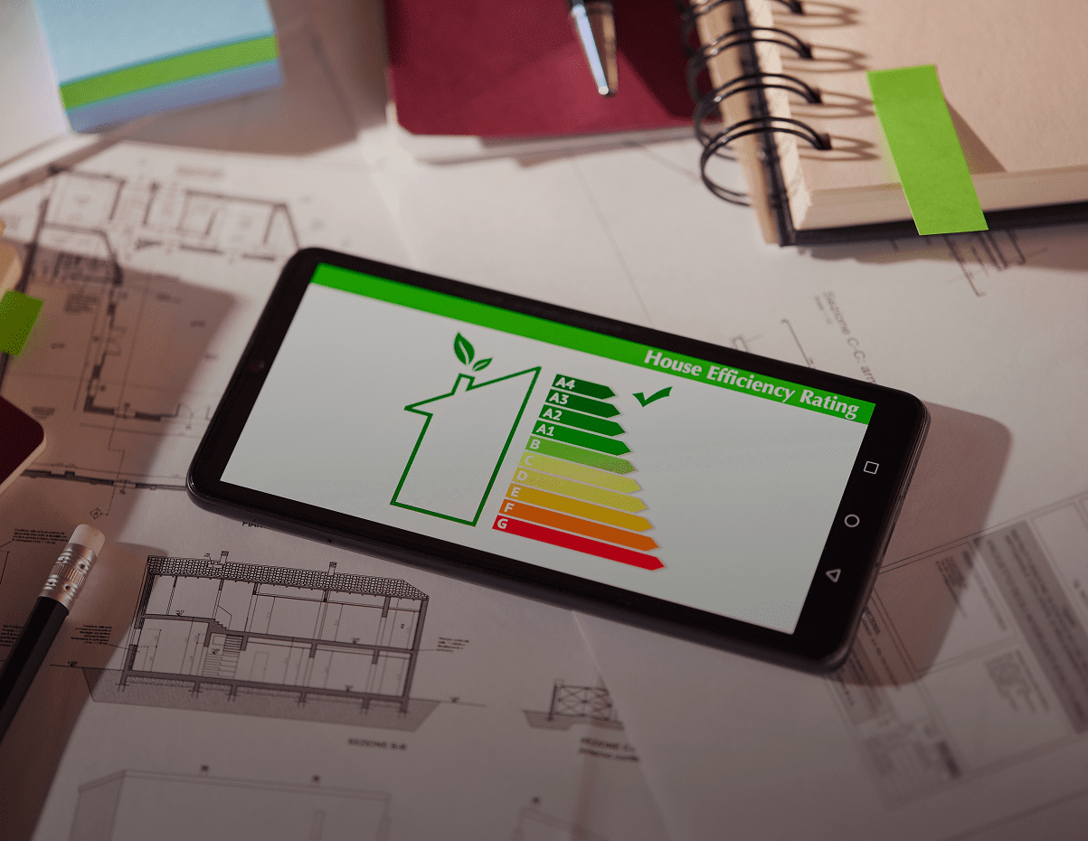 A smartphone on architectural plans displays a house efficiency rating chart with color bars from green (A) to red (G) and a green checkmark next to the top rating.
